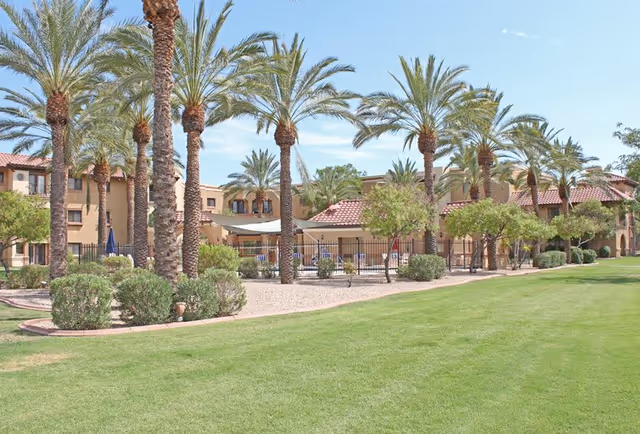 Exterior view of The Forum at Desert Harbor facility showing a well-maintained lawn, palm trees, shrubs, and a two-story building with a red-tiled roof under a clear blue sky.