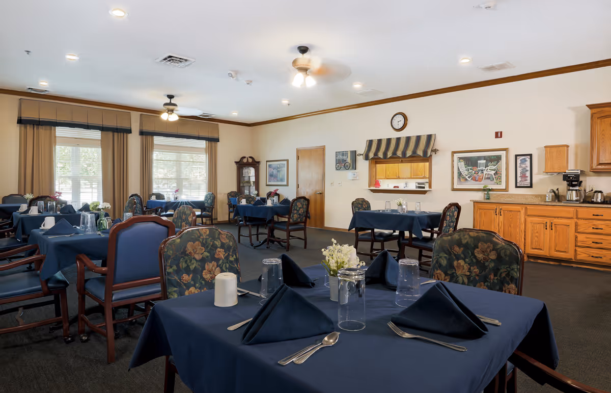 A dining room in an assisted living facility with multiple tables covered in dark blue tablecloths, each set with utensils, napkins, glasses, and small flower arrangements. The room has floral upholstered chairs, large windows with beige curtains, ceiling fans, and wooden cabinetry along one wall with a coffee station.