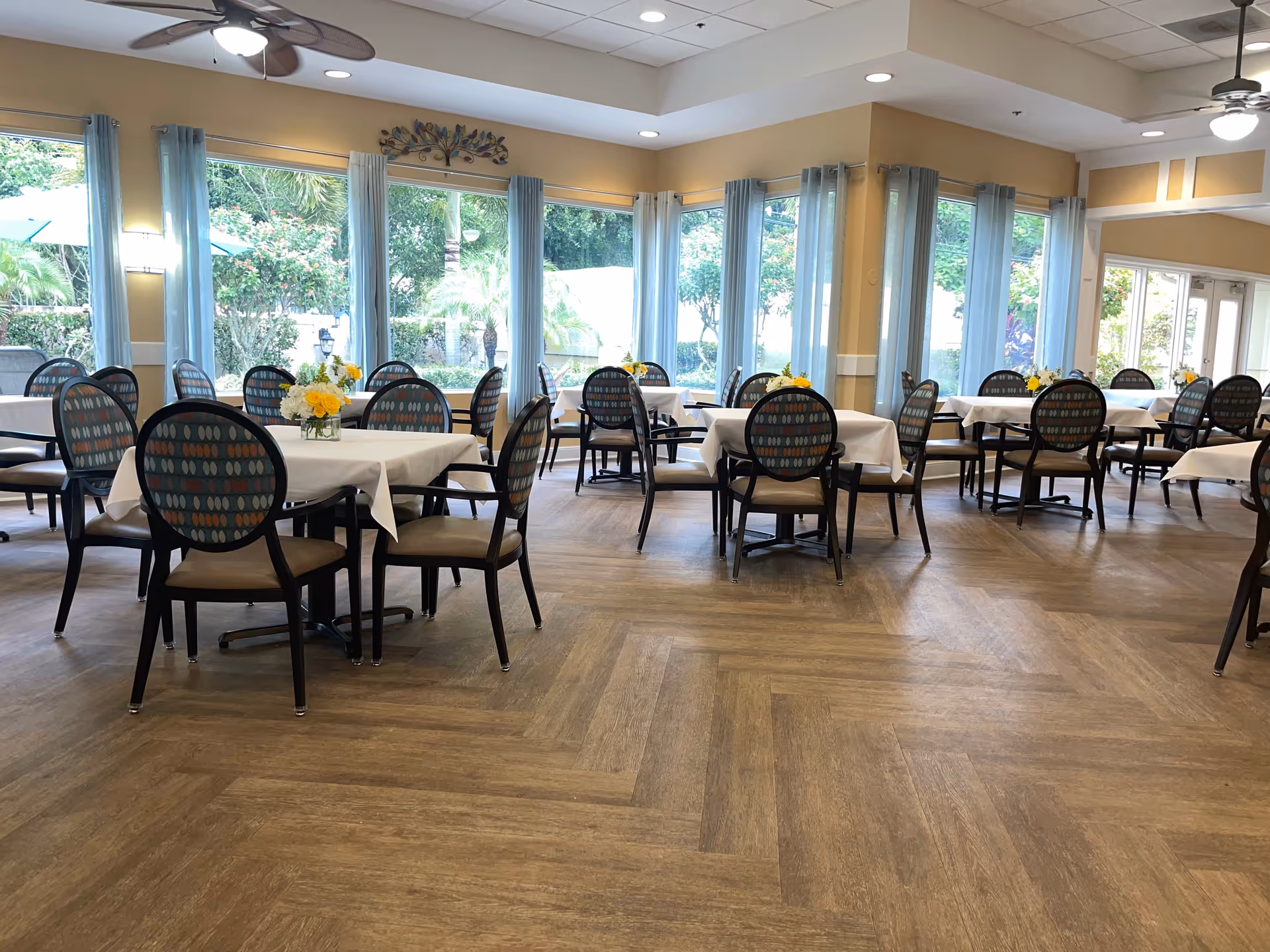 A bright dining room with multiple tables covered with white tablecloths, each surrounded by chairs with patterned backs. Small flower arrangements are placed on each table. Large windows with light blue curtains allow natural light to fill the room, and ceiling fans are visible above.