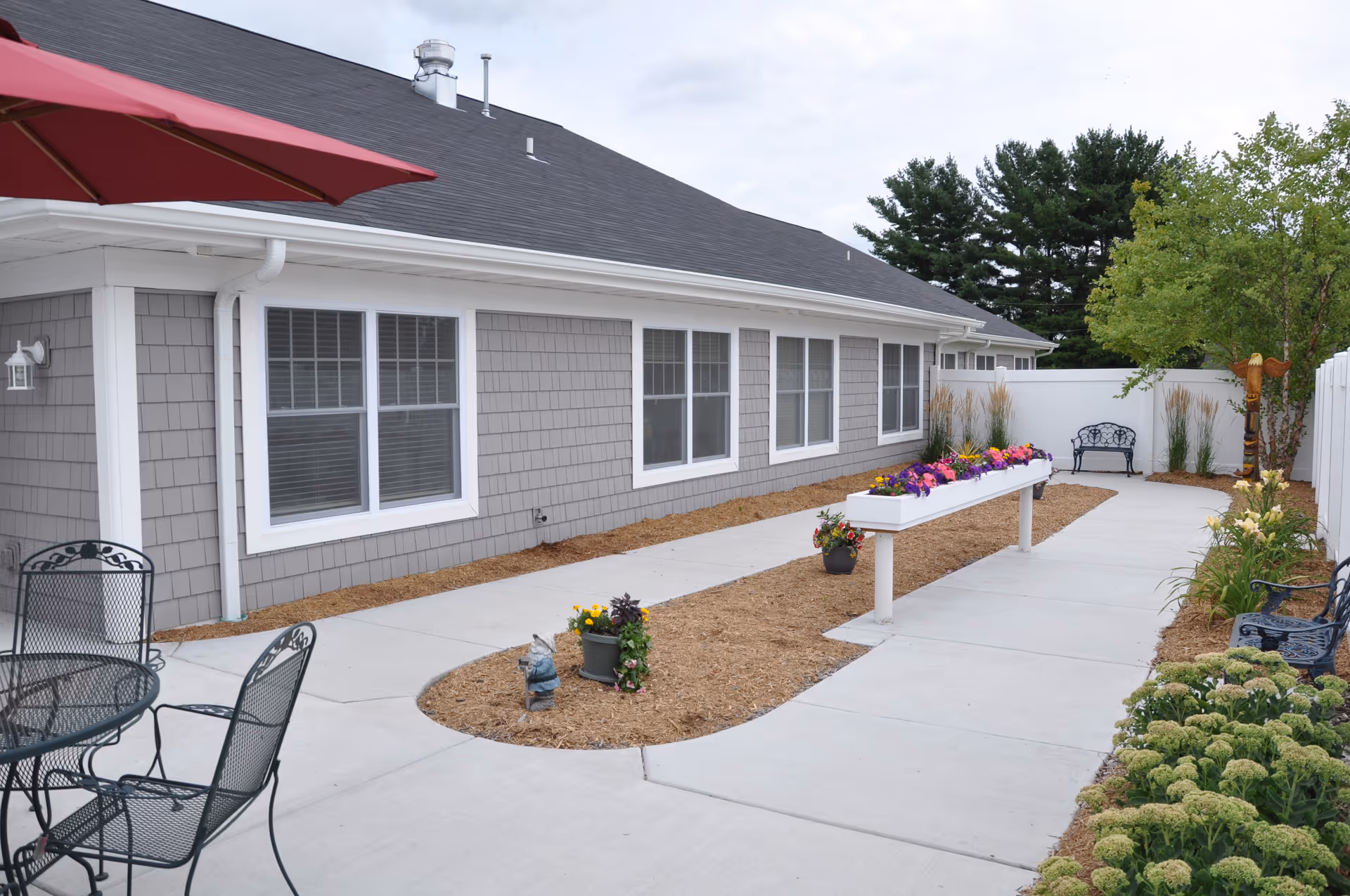 Outdoor patio area at Huntington Place featuring a concrete walkway, flower beds with colorful flowers, metal chairs and tables, a red umbrella, and a bench near a white fence with trees and shrubs in the background.