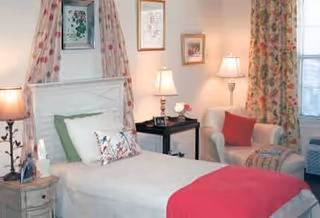 A cozy bedroom featuring a single bed with white bedding, green and floral pillows, and a red throw blanket. The room includes a white armchair with a red pillow, two bedside tables with lamps, floral curtains, and framed artwork on the walls.