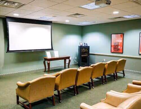 A small theater or presentation room with two rows of tan upholstered chairs facing a white projection screen mounted on a green wall. There is a small wooden table beneath the screen and a black cabinet with electronic equipment to the right. Two framed posters hang on the wall to the right.