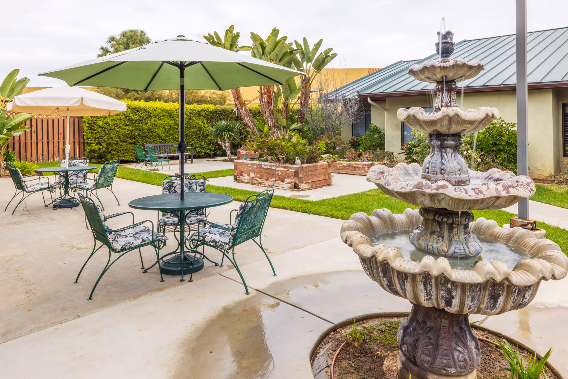 Outdoor patio area with green metal tables and chairs, some with umbrellas, surrounded by greenery and plants. A multi-tiered stone water fountain is in the foreground, with water flowing from the top tier. The background shows part of a building with a green roof and garden beds.