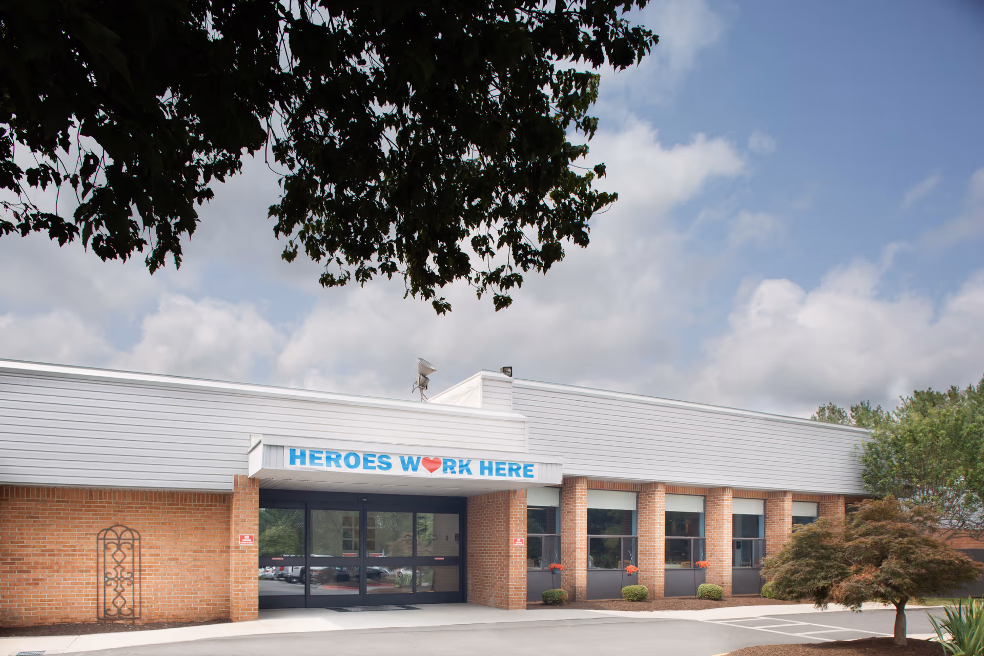 Exterior view of a single-story brick building with a white roof and large windows. Above the entrance, there is a sign that reads 'HEROES WORK HERE' with a red heart symbol. Trees and shrubs are visible around the building under a partly cloudy sky.