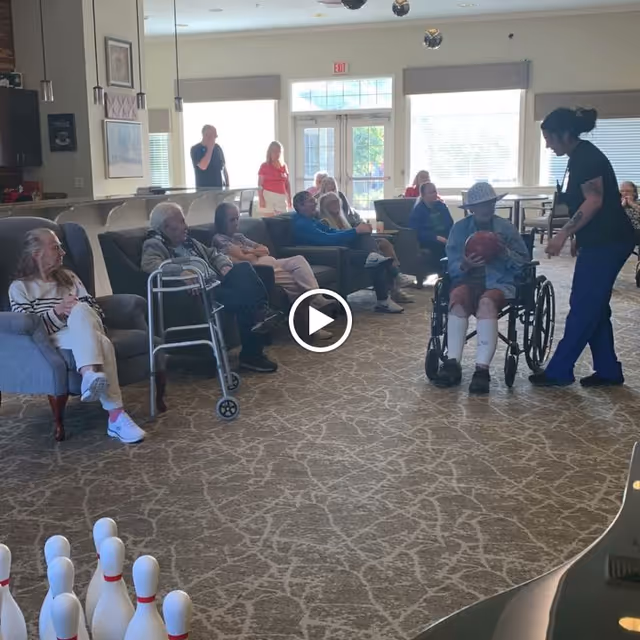 Residents and staff in a memory care common room playing a seated bowling game, with a wheelchair user holding a ball and others seated on sofas and chairs.