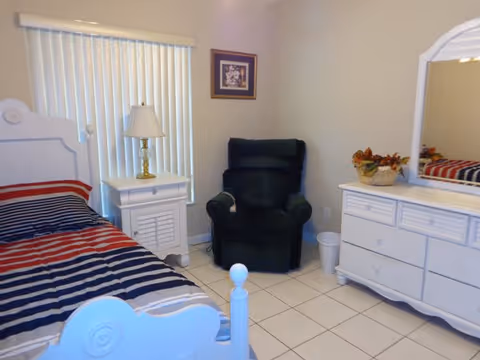 A bright bedroom with a white bed with striped bedding, a nightstand and lamp, a green recliner, and a white dresser with mirror.