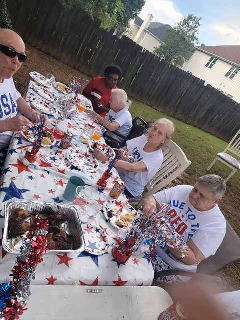 A group of elderly people sitting around a table outdoors, enjoying a meal together. The table is covered with a patriotic star-patterned tablecloth and decorated with red, white, and blue centerpieces. There are plates with food, cans of soda, and cups on the table. The setting is a fenced backyard with grass and trees in the background.