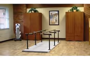 A rehabilitation room with parallel bars for physical therapy in the center, two wooden cabinets against a brick wall, a framed picture hanging between the cabinets, a window on the left side, and a medical device near the window.