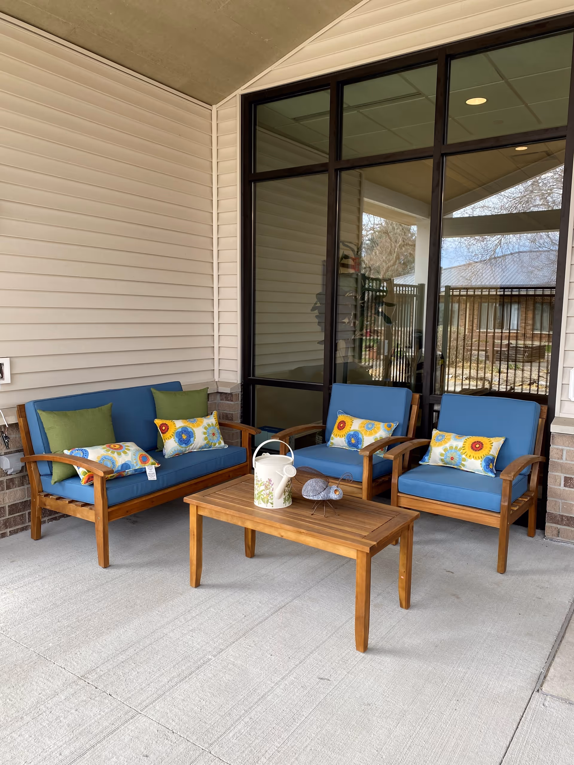 Outdoor seating area with a wooden loveseat and two wooden armchairs, all with blue cushions and colorful floral pillows. A wooden coffee table sits in front of the seating with a decorative watering can and a small metal bird sculpture on top. The area is covered and adjacent to a building with beige siding and large glass windows.