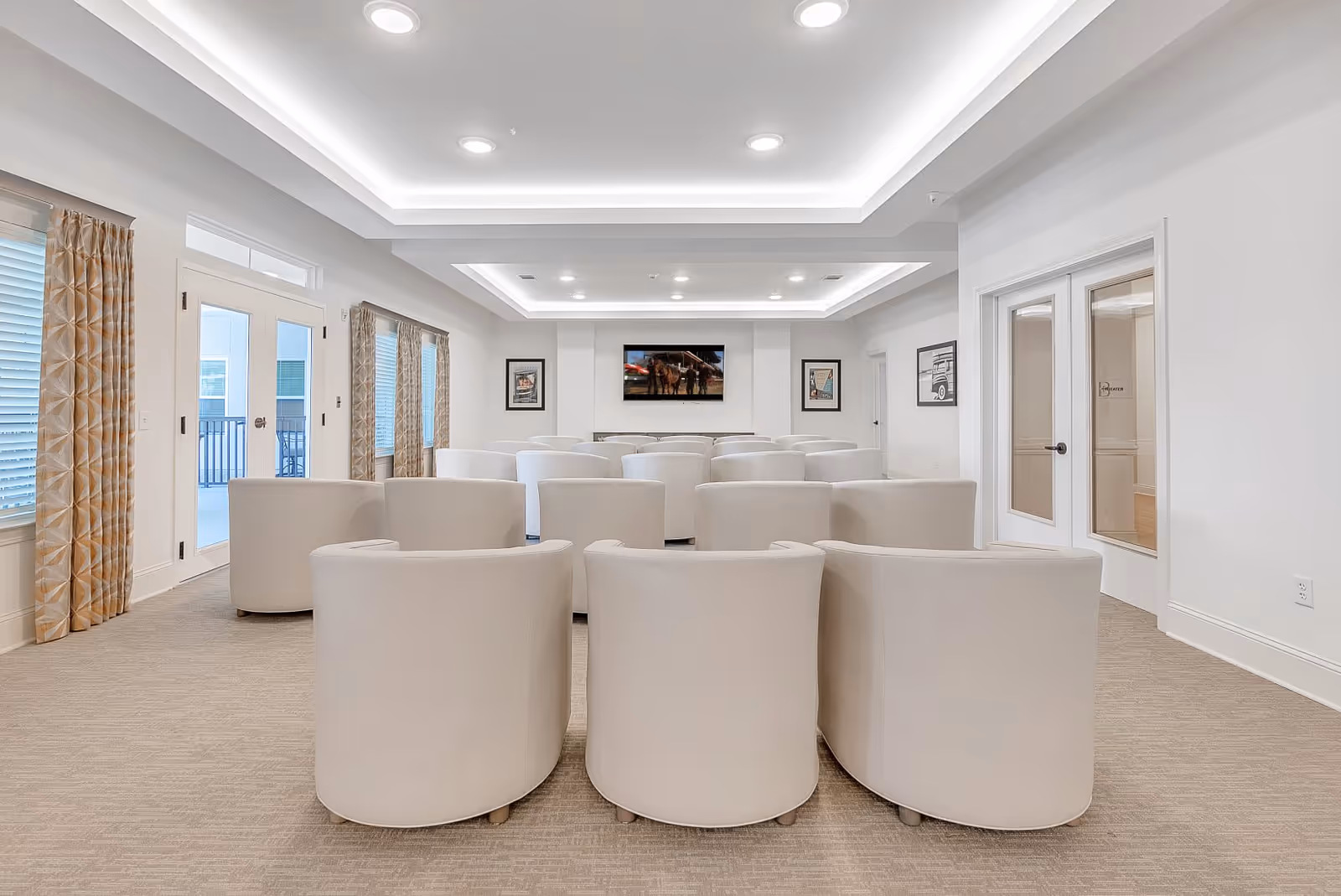A modern meeting or theater room with multiple rows of beige armchairs facing a wall-mounted flat screen TV. The room has white walls, recessed ceiling lights, and large windows with patterned curtains. Double glass doors are visible on the right side.