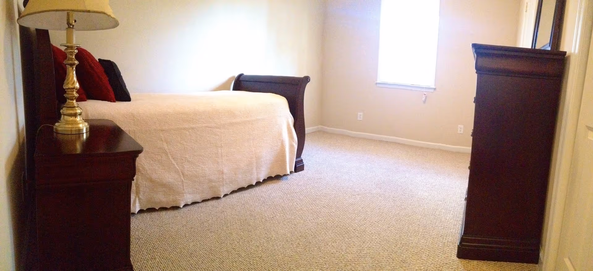 A bedroom with beige carpet and cream-colored walls. The room features a dark wooden bed with a white bedspread and red and black pillows. There is a matching dark wooden nightstand with a brass lamp on the left side of the bed and a tall dark wooden dresser on the right side near a window with closed blinds.