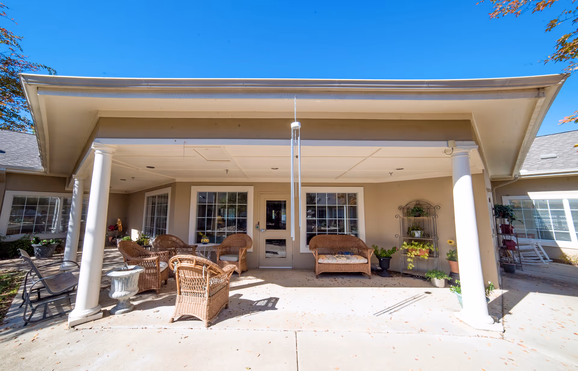 Covered outdoor patio area with wicker chairs, a wicker loveseat with a cushion, a small table, and various potted plants. The patio is supported by white columns and has a wind chime hanging from the ceiling. The background shows windows and a door leading inside the building under a clear blue sky.