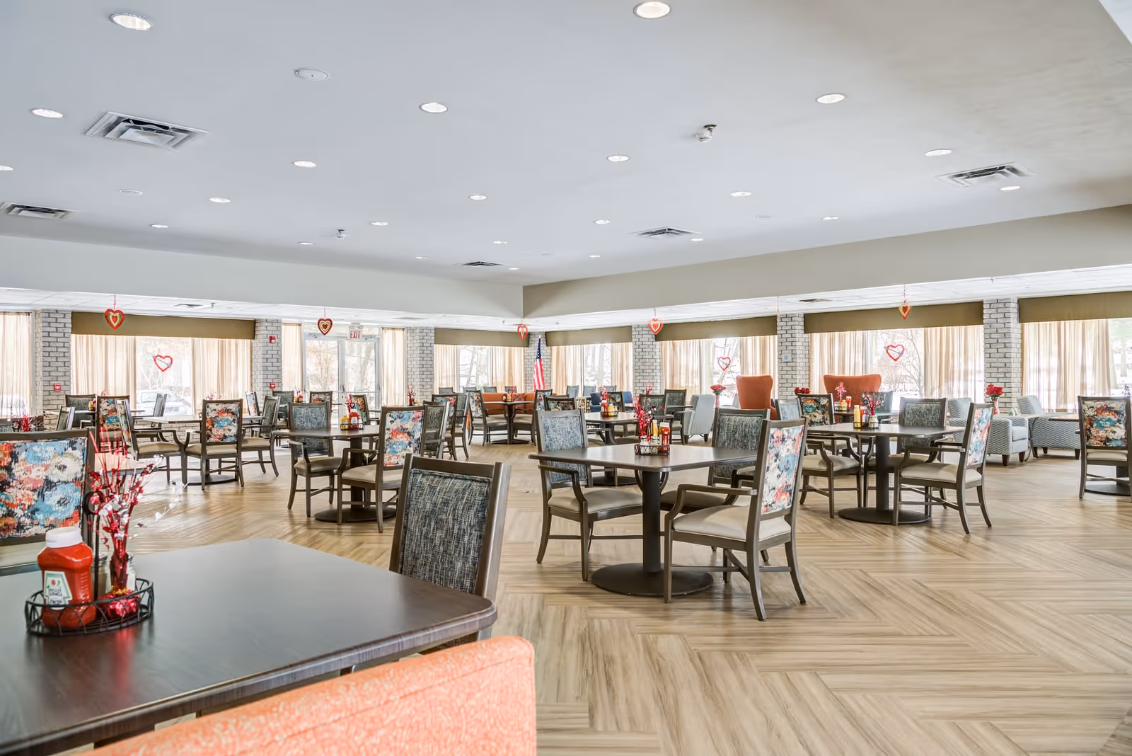 A spacious senior living dining room with multiple tables and chairs arranged neatly. The chairs have floral and patterned upholstery. The room has large windows with beige curtains, letting in natural light. Heart-shaped decorations hang from the ceiling and windows, suggesting a Valentine's Day theme. The floor has a light wood pattern, and there are condiment holders with ketchup bottles and decorative items on the tables.