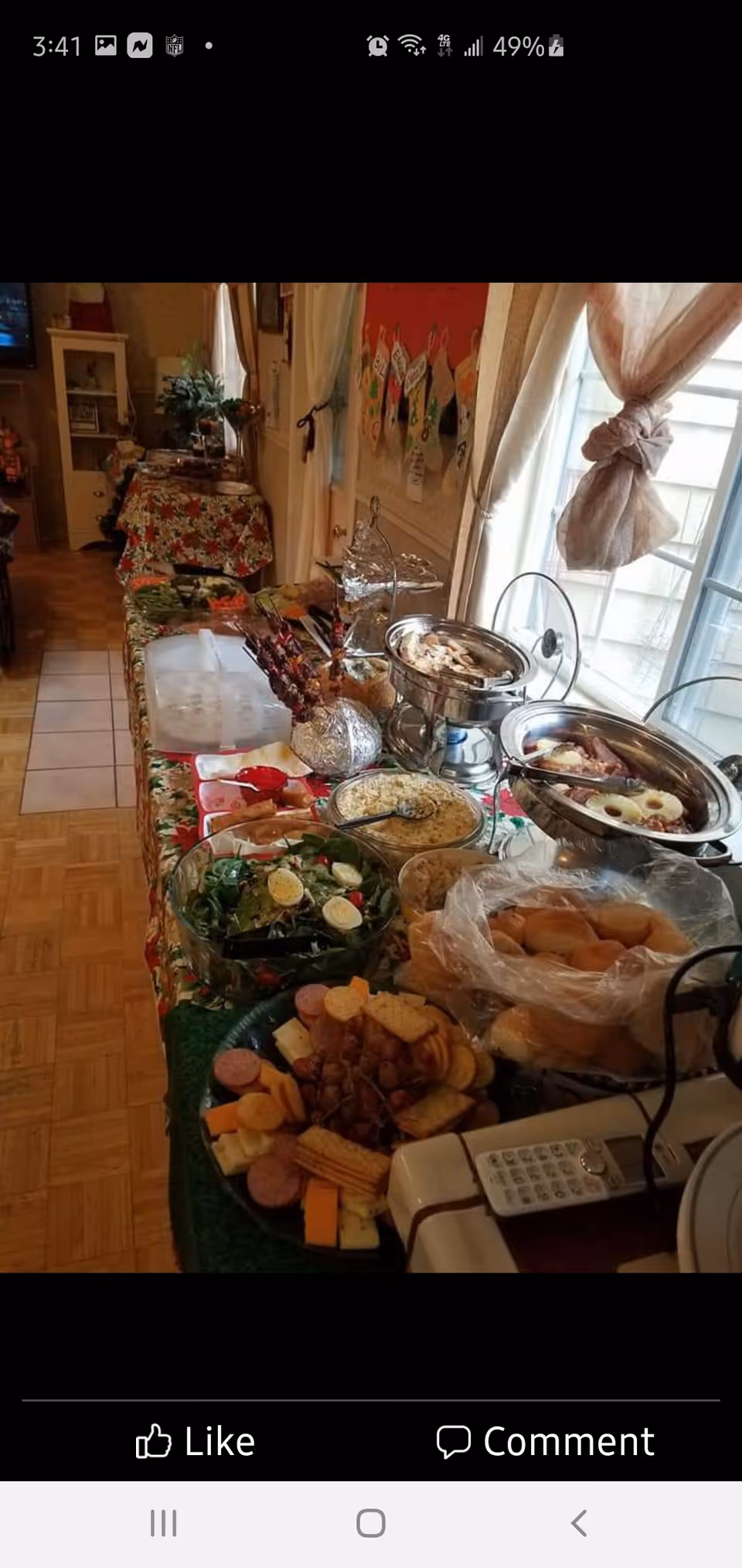 Buffet-style table in a dining area with chafing dishes, salads, rolls and assorted platters by a window with curtains.