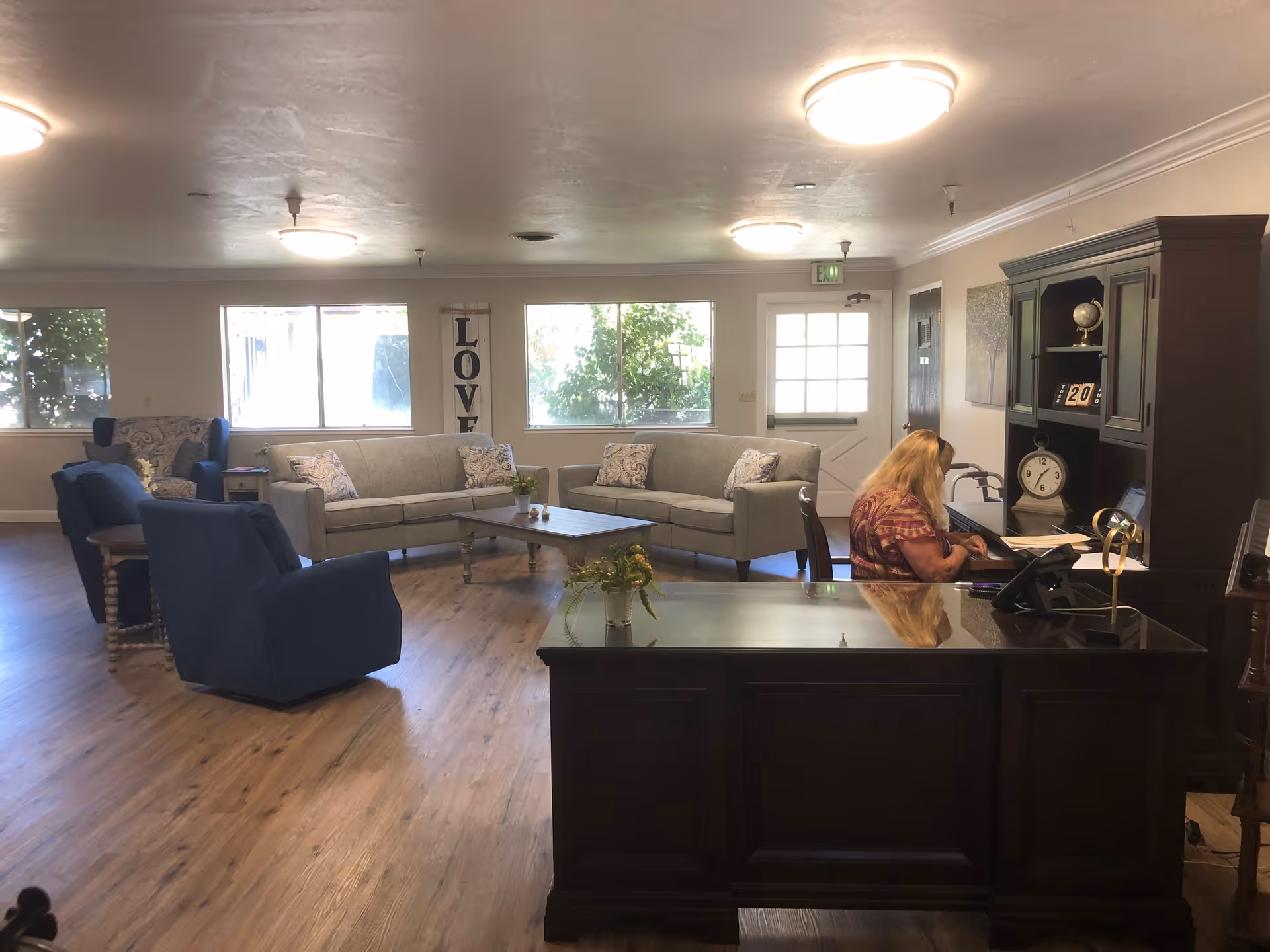 A cozy assisted living facility common area with wooden flooring, two gray sofas, two blue armchairs, a wooden coffee table, and a dark wooden desk with a woman sitting and working. Large windows let in natural light, and a vertical wall decoration with the word 'LOVE' is visible between the windows.