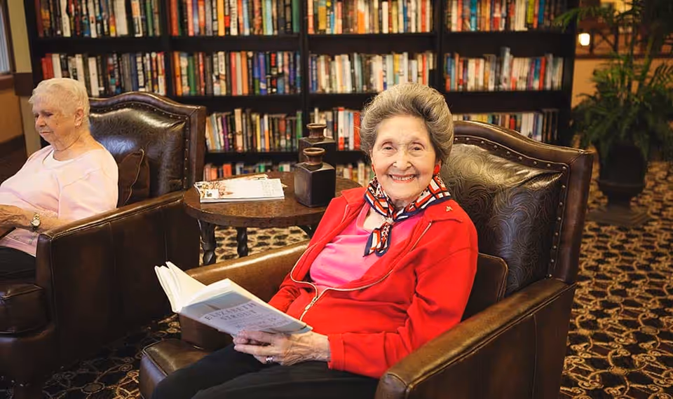 Two elderly women seated in leather armchairs in a cozy library/lounge area, one smiling and holding an open book in front of bookshelves.