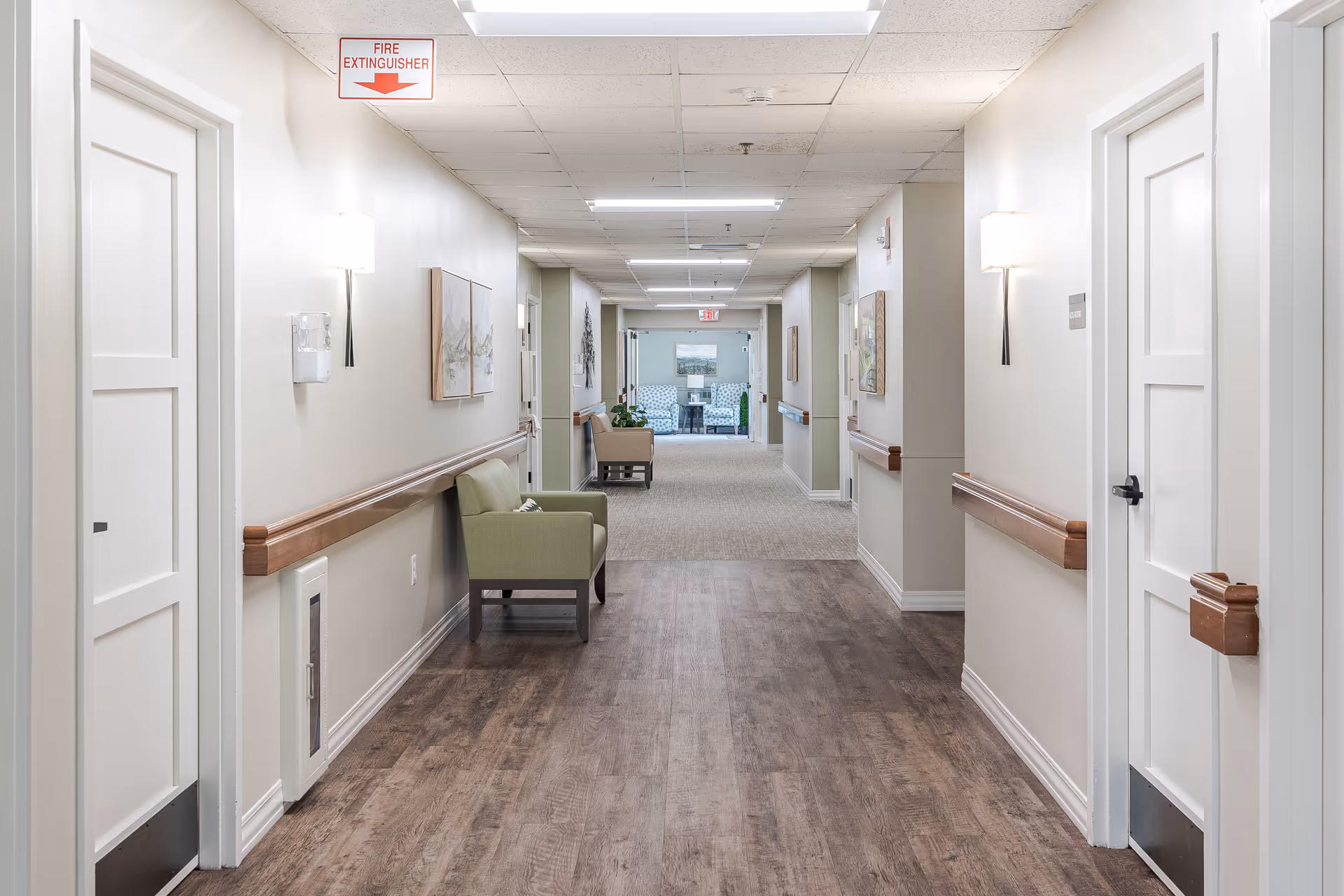 A well-lit hallway in a senior living facility with wood-look flooring and beige walls. The hallway features white doors on both sides, wooden handrails, wall-mounted lights, and framed artwork. At the far end, there is a seating area with two patterned armchairs and a small table, along with some plants. A fire extinguisher sign is visible on the ceiling.