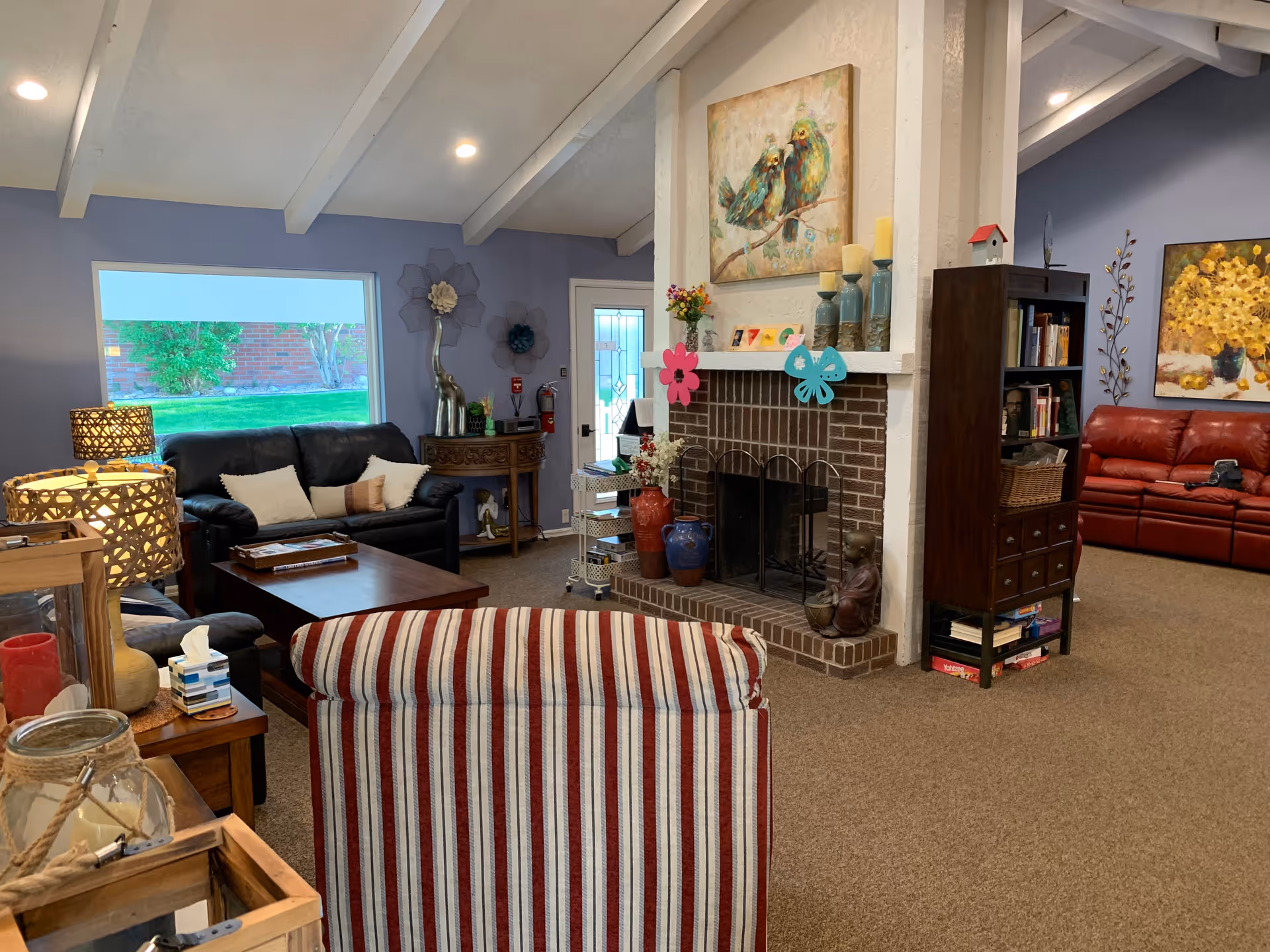 A cozy living room with a striped armchair in the foreground, a black leather sofa with pillows, a wooden coffee table, and a brick fireplace decorated with vases, candles, and colorful paper flowers. The walls are painted light blue, and there are various decorative items including paintings of birds and flowers, a bookshelf, and lamps. A window shows a green lawn outside.