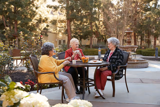 Three elderly women sitting around a round outdoor table in a garden area, enjoying drinks and snacks. They are engaged in conversation with trees, flowers, and a stone fountain visible in the background.