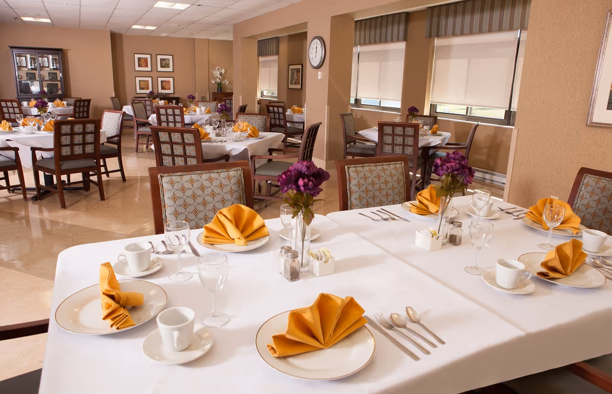 A well-lit dining room in a senior living facility with multiple tables set for a meal. Each table has white tablecloths, neatly folded yellow napkins, white plates, cups, glasses, and silverware. Purple flower centerpieces decorate the tables. The room has beige walls, large windows with blinds, framed artwork, and a clock on the wall.