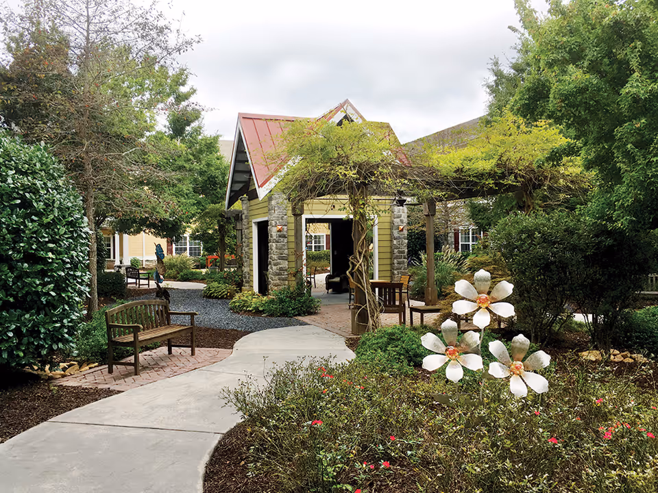 Outdoor garden area at Cobblestone At Park Springs featuring a paved walkway, wooden benches, lush green trees and shrubs, a small stone and wood pavilion with a red roof, and decorative metal flowers in the foreground.