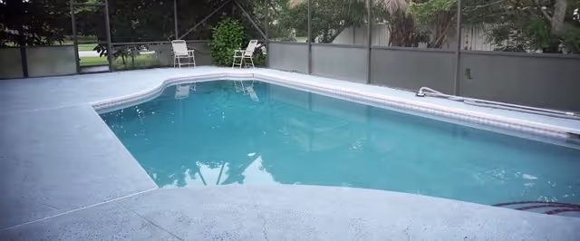 An outdoor swimming pool with clear blue water surrounded by a concrete deck. Two white chairs are placed near the far end of the pool, and the area is enclosed by a screened fence with greenery visible outside.