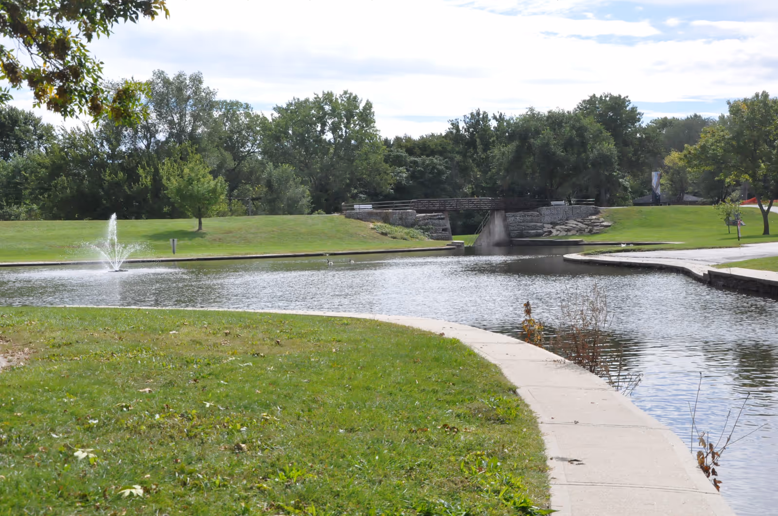 Park pond with a small fountain, curving concrete walkway, grassy banks, and a pedestrian bridge in the background.