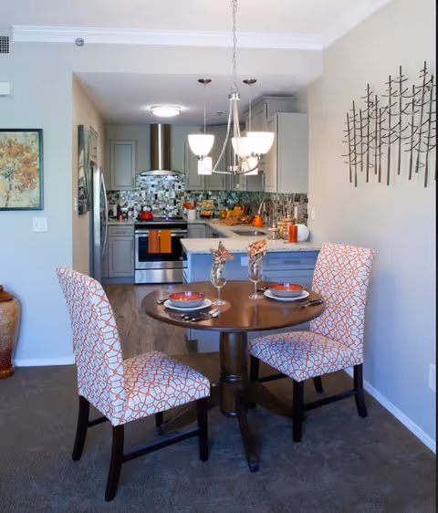 A cozy dining area with a round wooden table set for two with plates, glasses, and napkins. Two upholstered chairs with a white and orange geometric pattern surround the table. In the background, there is a modern kitchen with gray cabinets, a stainless steel refrigerator, stove, and a mosaic tile backsplash. A chandelier with five lights hangs above the dining table, and decorative wall art is visible on the right wall.