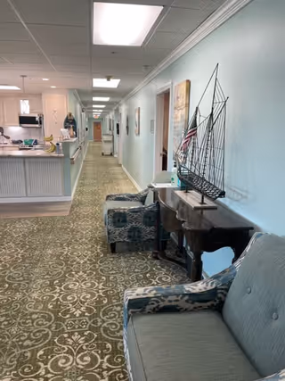 A long hallway in a senior living facility with light blue walls and patterned green carpet. On the right side, there is a wooden table with a decorative metal sailboat and an American flag, along with two upholstered chairs. On the left side, there is a reception or kitchen counter area with white cabinetry and a microwave. The hallway is well-lit with ceiling lights and has several doors and framed pictures along the walls.