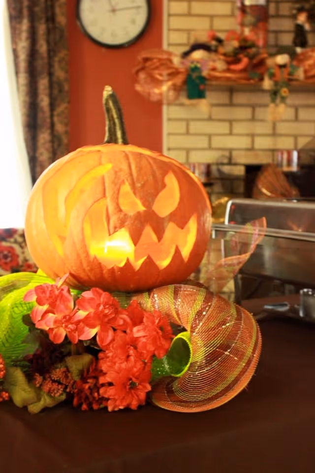 Carved glowing jack-o'-lantern displayed on a table centerpiece with red flowers and decorative ribbon in a cozy indoor room.