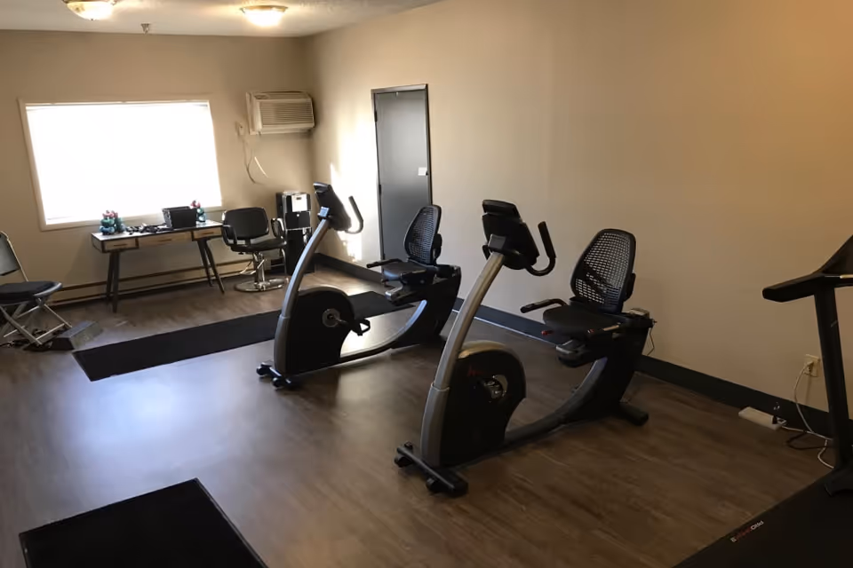 A small exercise room with two recumbent stationary bikes, a treadmill, and a desk with chairs near a window. The room has wood flooring and beige walls with a wall-mounted air conditioning unit.