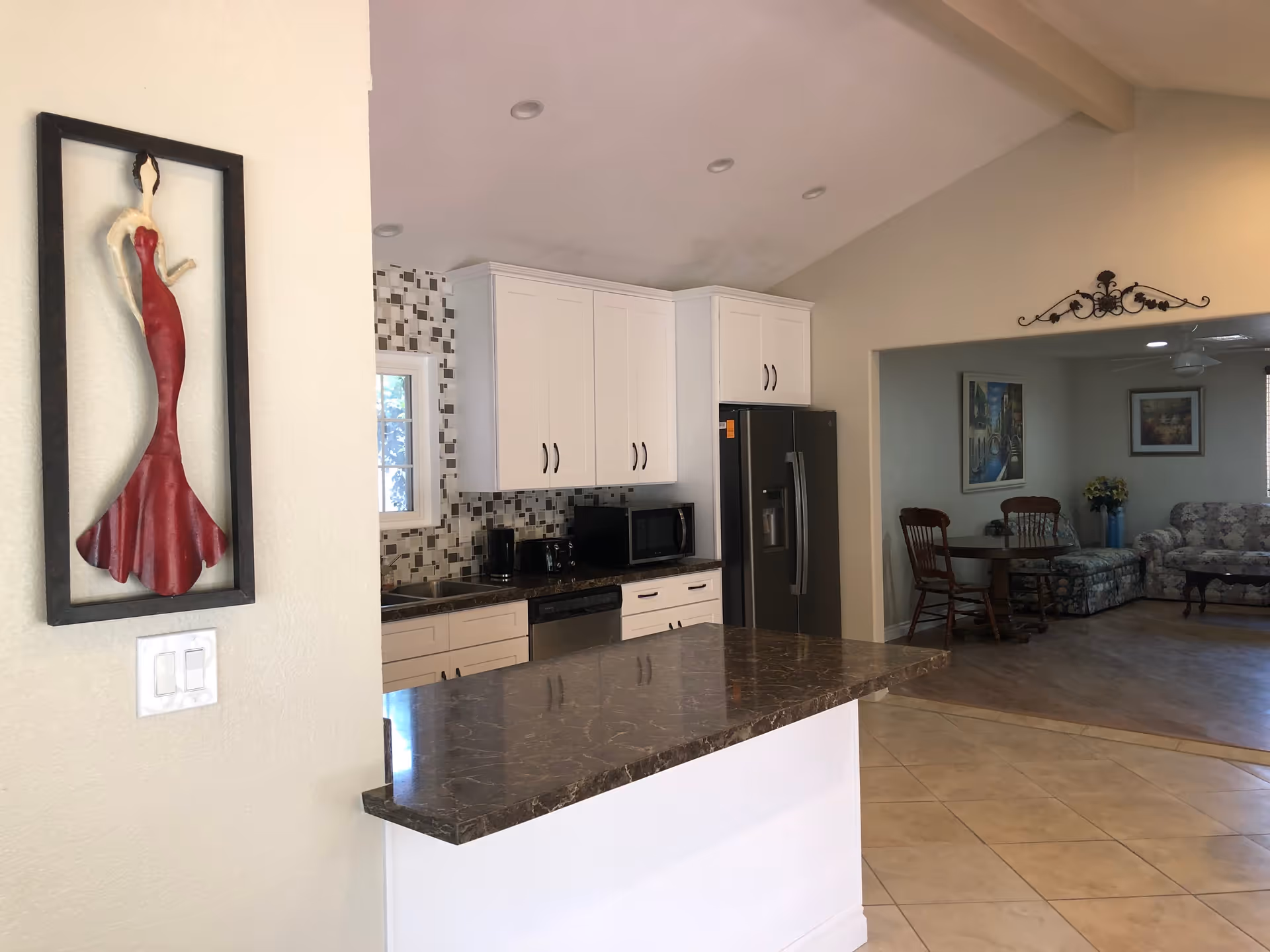 Interior view of a senior living facility showing a kitchen with white cabinets, a dark marble countertop, a black refrigerator, microwave, toaster, and coffee maker. Adjacent to the kitchen is a living room area with a round wooden table, chairs, a floral patterned couch, and framed artwork on the walls. A decorative wall sculpture of a woman in a red dress is mounted on a nearby wall.