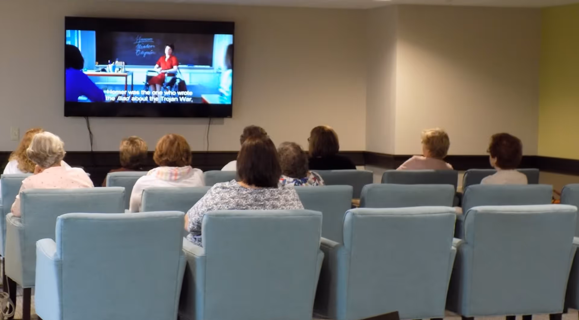 A group of elderly people seated in light blue armchairs facing a wall-mounted television screen in a common room, watching a program with subtitles.