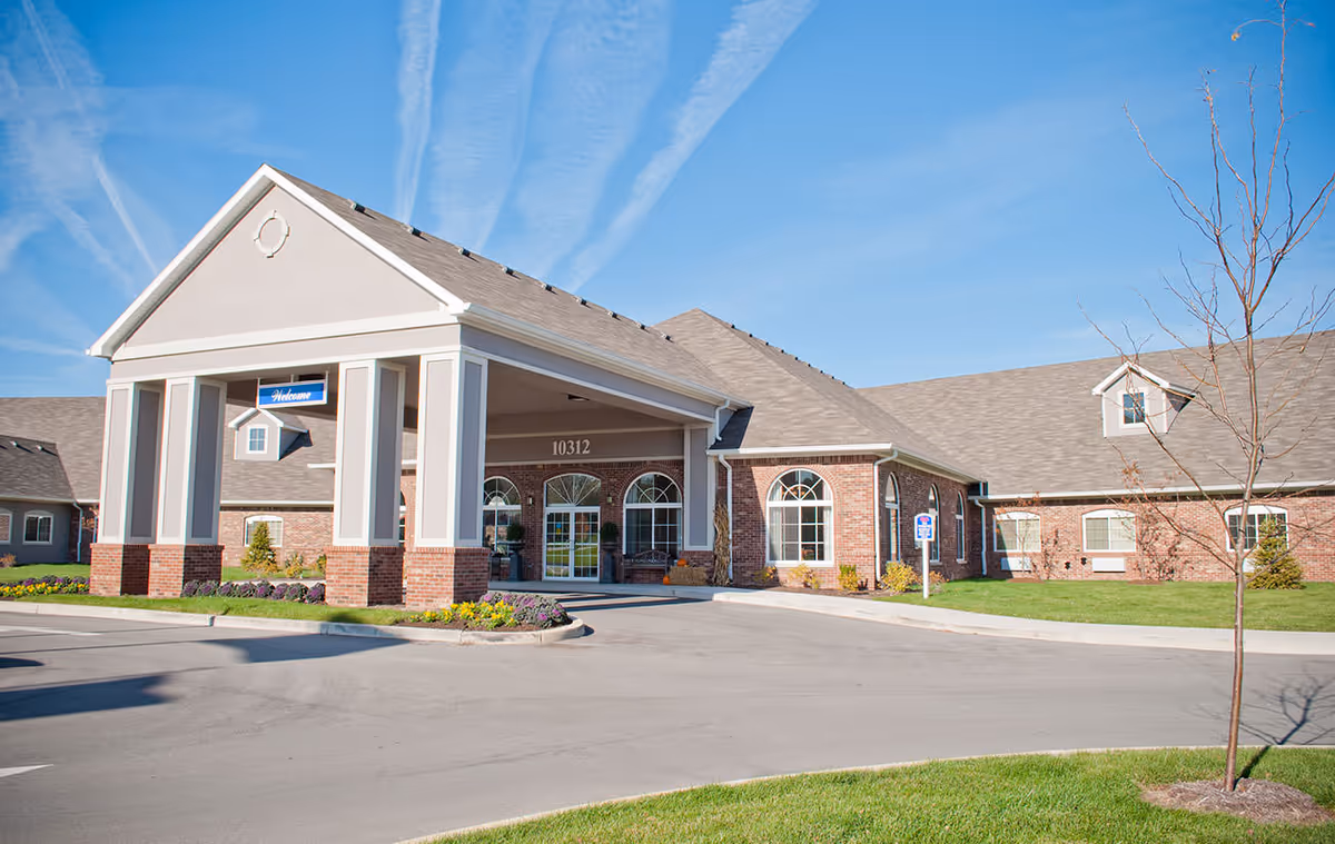 Exterior view of Allisonville Meadows facility showing a brick building with a covered entrance, large windows, and a clear blue sky with contrails above.