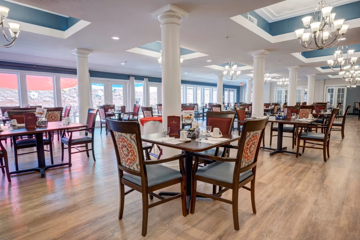 A spacious dining room with multiple wooden tables and chairs, each table set with placemats, cups, and menus. The room features large windows allowing natural light, white columns, chandeliers, and a wooden floor.