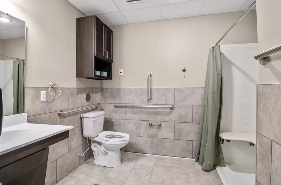 A clean and accessible bathroom featuring a white toilet with grab bars on the walls around it, a white sink with a dark wood vanity, a wall-mounted dark wood cabinet with towels, and a shower area with a green curtain and a fold-down seat.