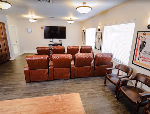 A bright common room with a row of leather recliner seats facing a wall-mounted TV, wood floors, and additional side chairs by the windows.