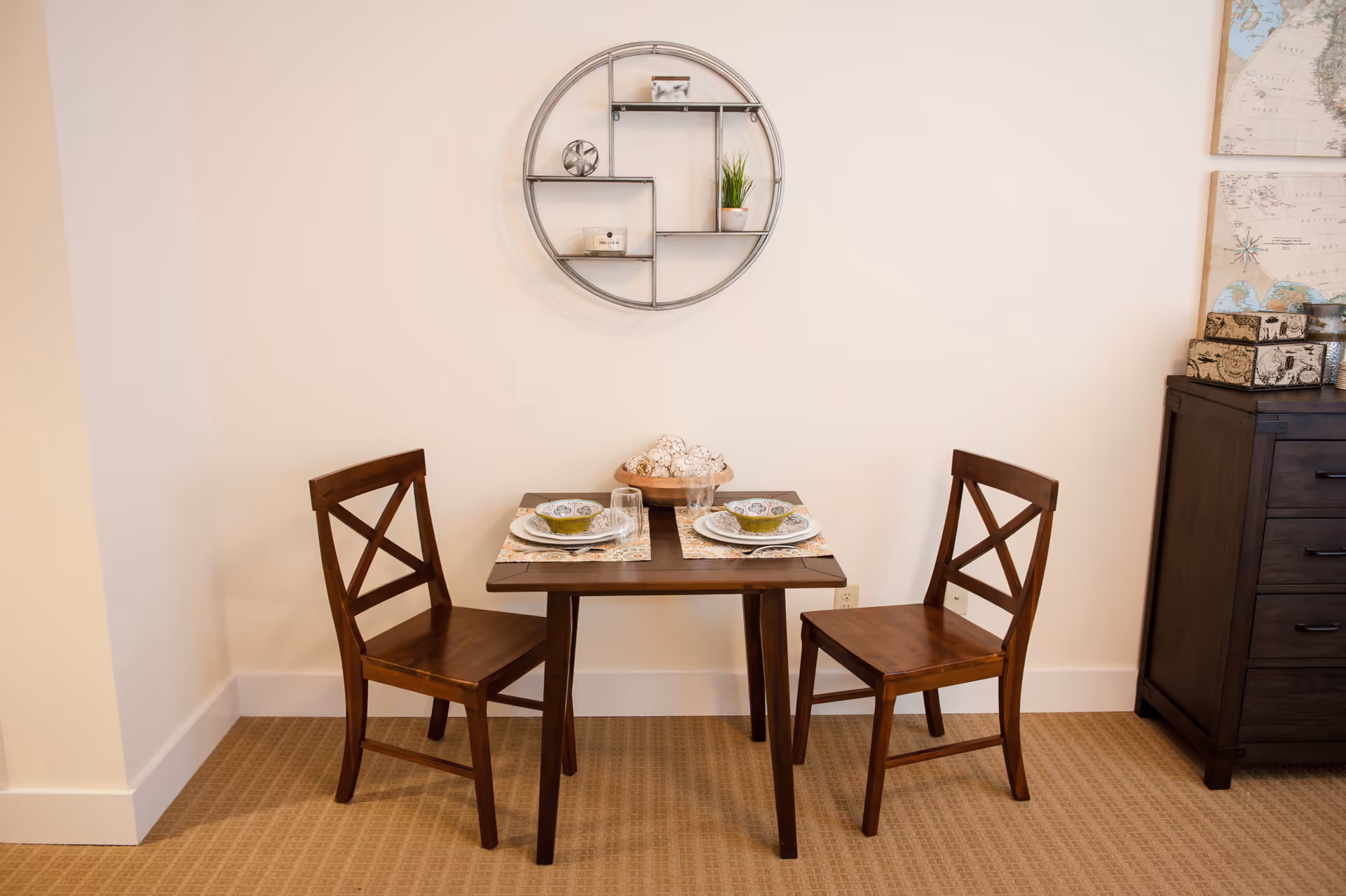 Small dining area with a wooden table set for two, two wooden chairs, a decorative round wall shelf and a dresser against a cream wall.