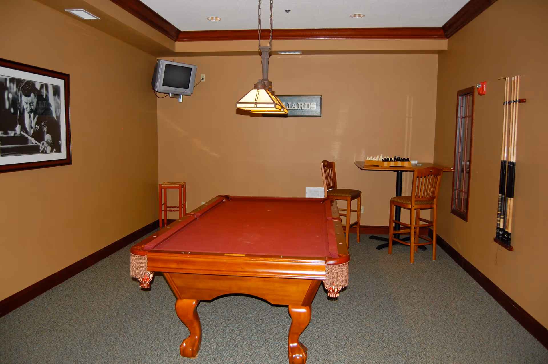 A game room with a red felt pool table in the center, a hanging light fixture above it, a small wall-mounted TV in the corner, a framed black and white photo of a man playing pool on the left wall, and a small table with two chairs and a chess set on the right side. Pool cues are mounted on the right wall.