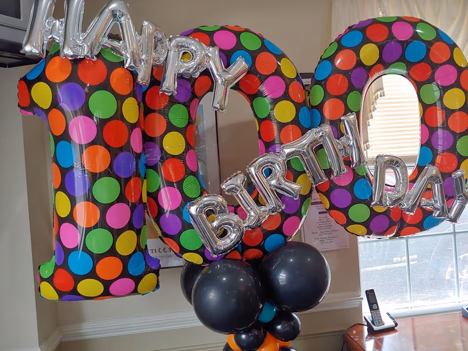 Colorful polka dot balloons shaped as the number 100 with silver letter balloons spelling out 'HAPPY BIRTHDAY' attached to a cluster of black and orange balloons, set indoors near a window with blinds and a wooden table holding a cordless phone.