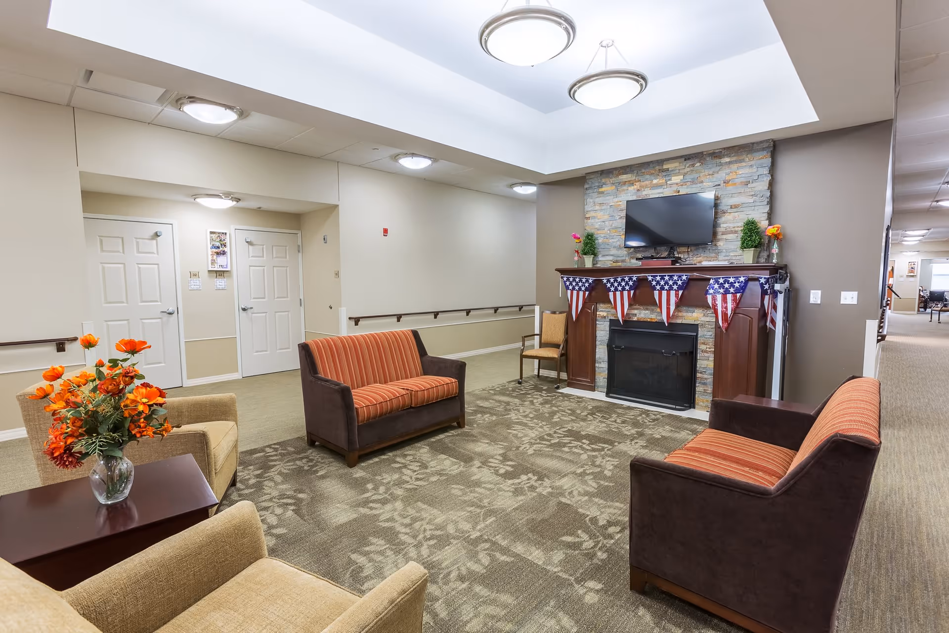 A cozy common area in a senior living facility with two brown and orange striped loveseats, two beige armchairs, a small wooden table with a vase of orange flowers, and a fireplace decorated with American flag bunting. A flat-screen TV is mounted above the fireplace, and the room has soft overhead lighting and beige walls.
