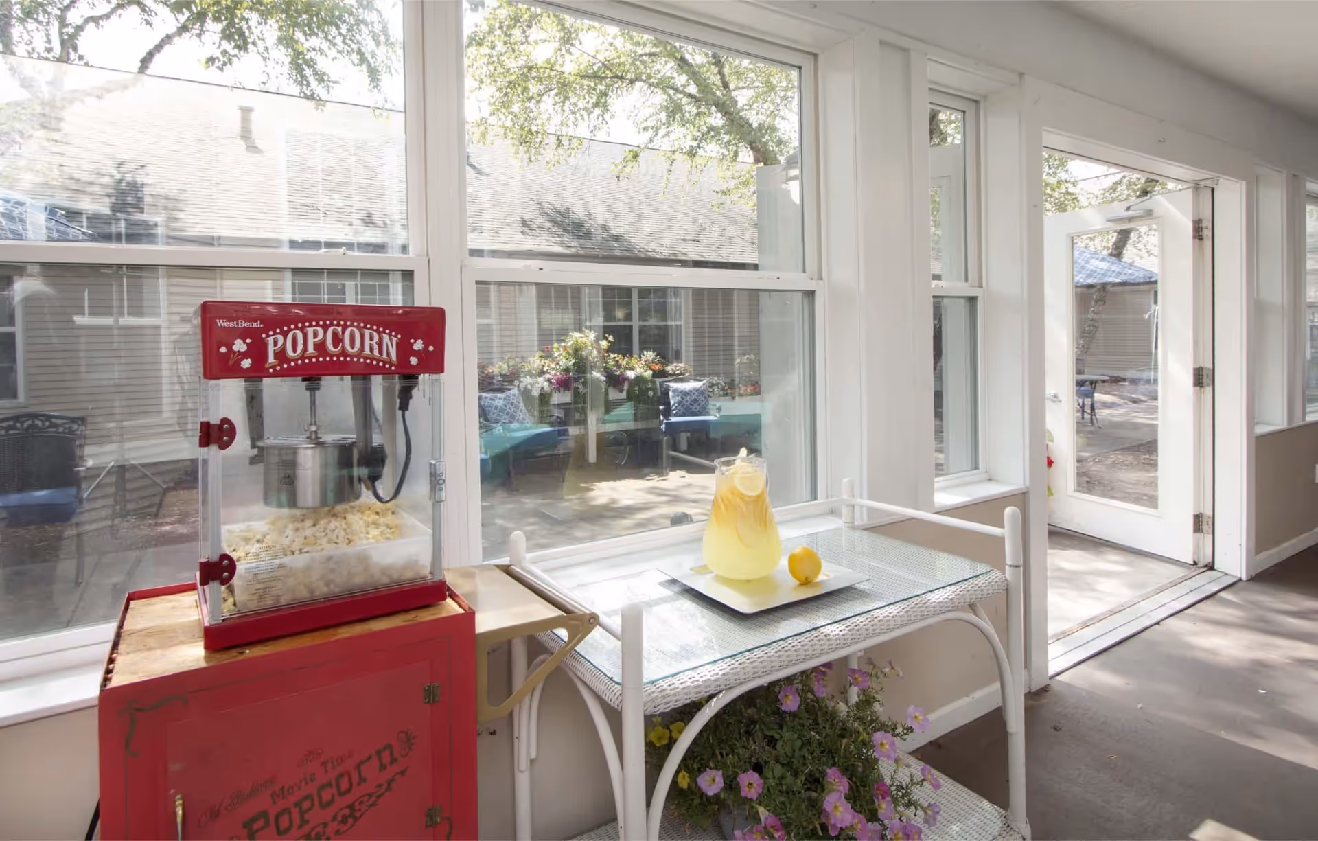A bright indoor area with large windows showing an outdoor patio. Inside, there is a red popcorn machine on a red cabinet to the left and a glass-top table with a pitcher of lemonade and a lemon on a tray. Below the table, there is a planter with purple flowers. The door to the patio is open, letting in natural light.