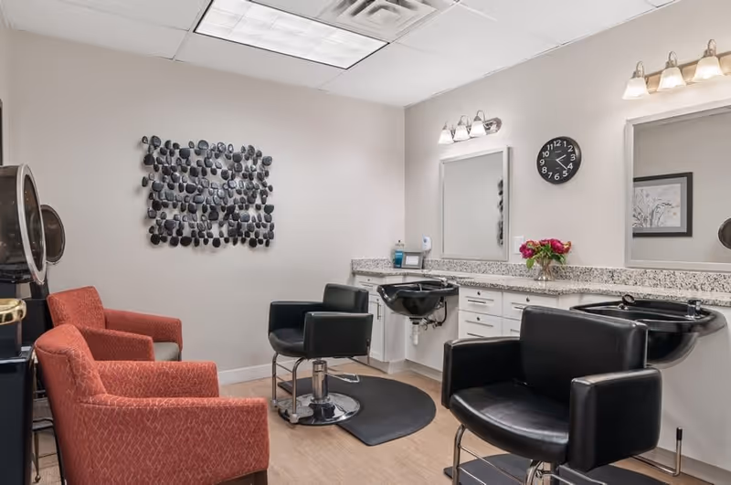 A salon room with styling chairs, shampoo sinks, mirrors, and red armchairs in a senior living facility.