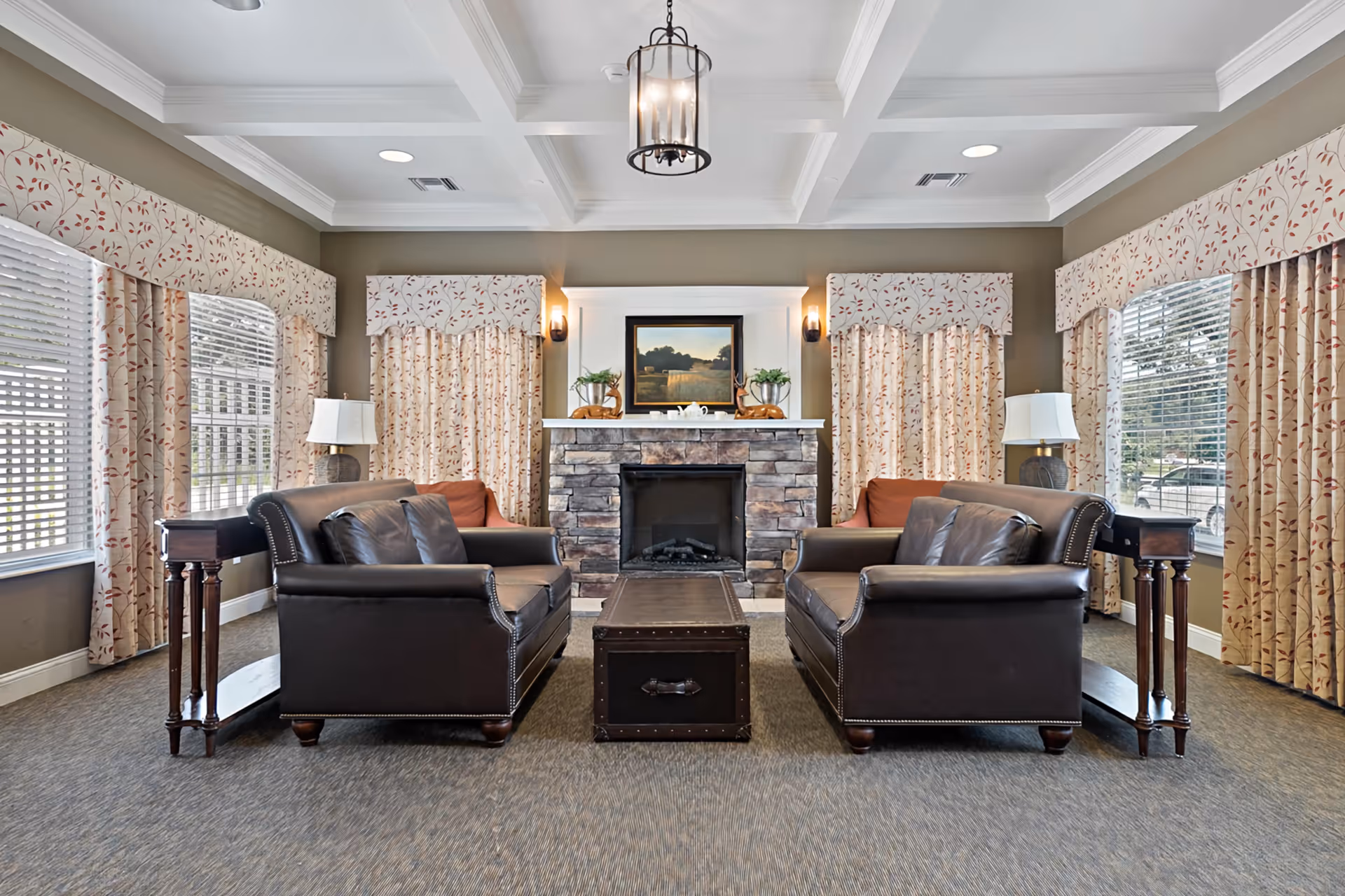 A cozy living room with two dark leather sofas facing each other across a wooden chest coffee table. The room features a stone fireplace with a framed landscape painting above it, flanked by two small plants and decorative items. Large windows with floral-patterned curtains allow natural light to fill the space. Two side tables with lamps are positioned beside each sofa, and a ceiling light fixture hangs above.