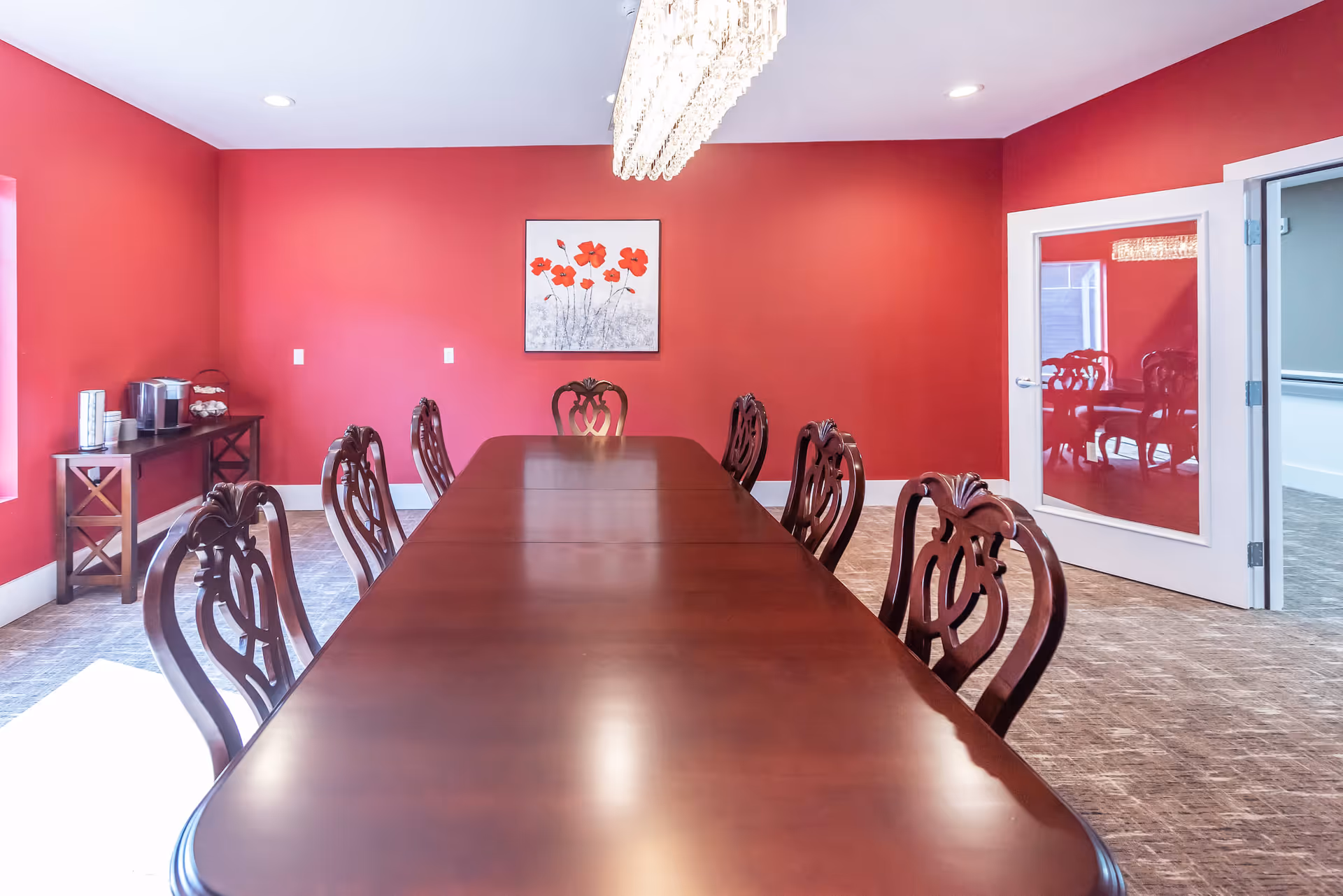 A formal dining room with a long wooden table surrounded by ornate wooden chairs. The walls are painted red, and there is a painting of red flowers on the far wall. A modern chandelier hangs above the table. To the left, there is a small table with a coffee maker and cups. A glass door on the right reflects part of the room.