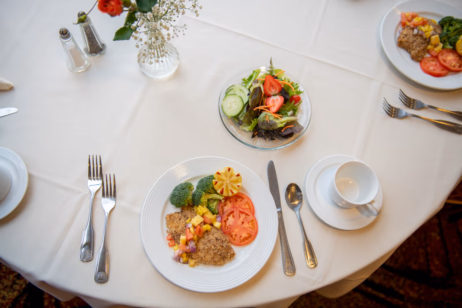 A dining table set with plates of food including breaded chicken with pineapple salsa, broccoli, tomato slices, and a side salad with strawberries, cucumber, and mixed greens. The table is covered with a white tablecloth and has silverware, a white cup and saucer, salt and pepper shakers, and a small vase with flowers.