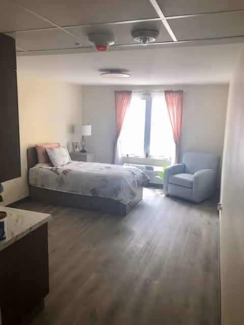 A bright senior living bedroom with a single bed, nightstand, armchair, and a window with pink curtains.