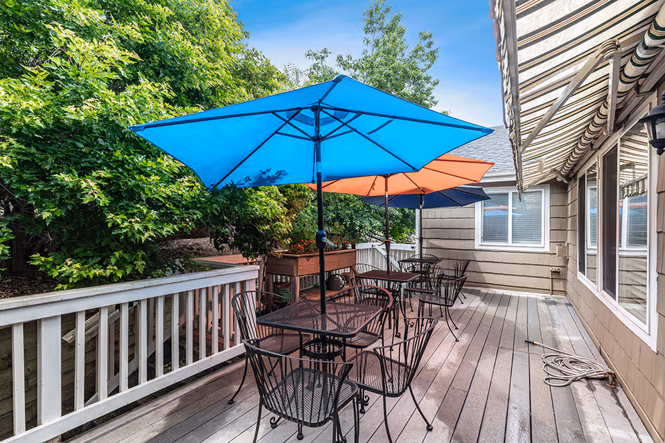 Outdoor patio area with metal tables and chairs under colorful umbrellas including blue, orange, and dark blue. The patio has a wooden deck and railing, with green trees and shrubs in the background. The side of a building with windows and striped awnings is visible on the right.