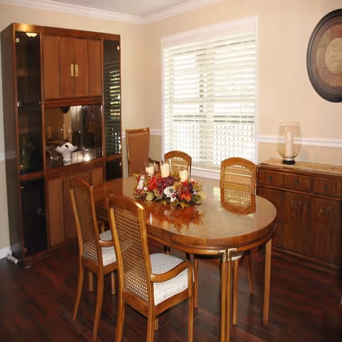A dining room with a wooden oval table surrounded by six wooden chairs with woven backs and cushioned seats. The table is decorated with a centerpiece featuring candles and flowers. There is a wooden cabinet with glass doors on the left side and a wooden sideboard with a decorative glass lamp on the right. A window with white blinds is centered on the back wall, and a round decorative wall piece hangs above the sideboard.