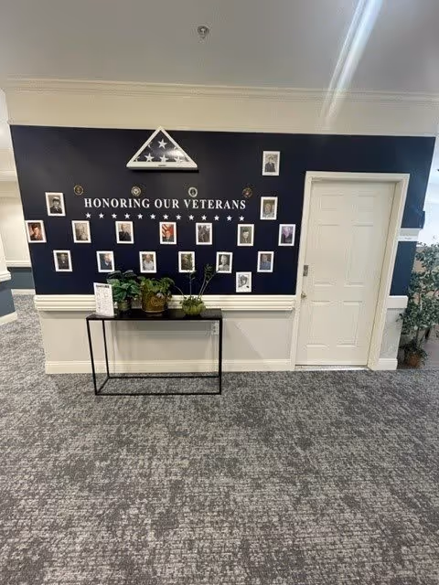 Interior wall display honoring veterans with framed photos and a folded American flag in a triangular case mounted above. Below the display is a black table with potted plants. The wall is painted dark blue with white trim, and there is a closed white door to the right.